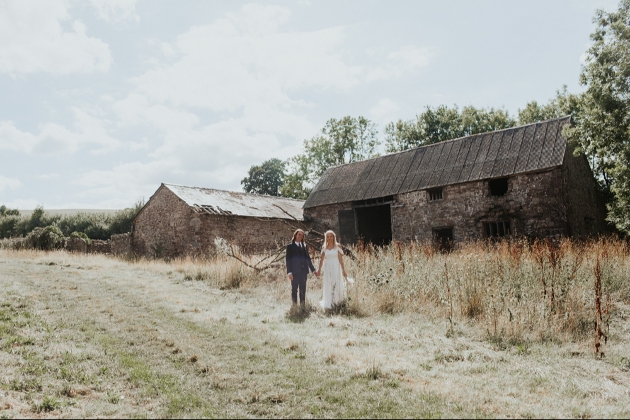 A bride and groom walking in a field in front of two barns