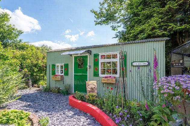 A small green shepherd's hut surrounded by trees and flowers