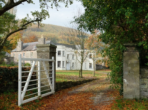 The exterior of a large white house surrounded by woodland with a gate in front