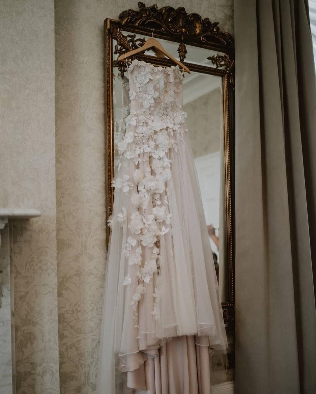 A blush, floral dress hanging on a hanger from a mirror