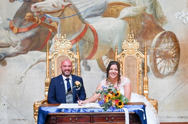 A bride and groom sitting on gold frames while signing a wedding registry